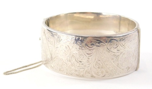 A 20thC silver hinged bangle, with heart engraved design of scrolls and vines, Birmingham 1949, engraved MBT, with safety chain, 6cm diameter, 47.2g all in.