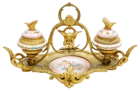 A 19thC French porcelain and ormolu mounted desk stand, possibly Sevres, the central dish painted with a cherub against a pink ground, flanked by two ink pots each cast with bird finials holding a letter in each beak, the porcelain ink pots decorated with