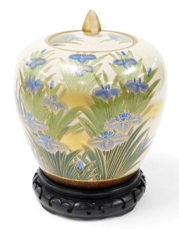 A Japanese earthenware jar and cover, decorated with iris' in blue, surrounded by leaves picked out in gilt, signed to underside and with silvered label 'Made In Japan', (AF), on a carved and ebonised stand, 25cm high.