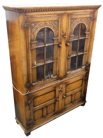 A Titchmarsh and Goodwin oak cabinet, the top with a moulded cornice and dentil frieze and two panelled and glazed doors, each with carved arches and brass hinges, the base with two panelled doors on stiles, 168cm high, 122cm wide.