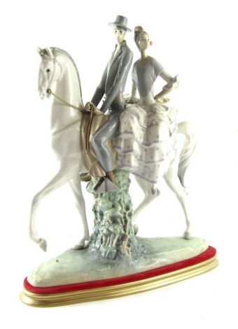 A Lladro porcelain figure group of a lady and gentleman riding on horseback, the gentleman in riding suit, with lady in a polka dot dress, mounted on a white horse, printed and impressed marks, on a gold and red plinth, 48cm high, 37cm wide. (AF)