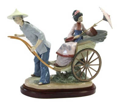 A Lladro porcelain figure group, depicting a Japanese gentleman in blue dress pulling a rickshaw carrying a lady with a parasol, raised on an oval wooden base, No 1383, 32cm high, 36cm wide.