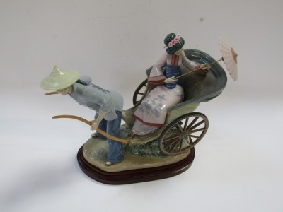 A Lladro porcelain figure group, depicting a Japanese gentleman in blue dress pulling a rickshaw carrying a lady with a parasol, raised on an oval wooden base, No 1383, 32cm high, 36cm wide. - 2