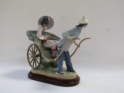 A Lladro porcelain figure group, depicting a Japanese gentleman in blue dress pulling a rickshaw carrying a lady with a parasol, raised on an oval wooden base, No 1383, 32cm high, 36cm wide. - 3