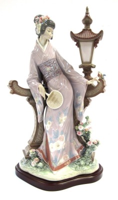 A Lladro porcelain figure of a Japanese geisha girl, modelled standing against a garden lantern, on a wooden base, 42cm high.