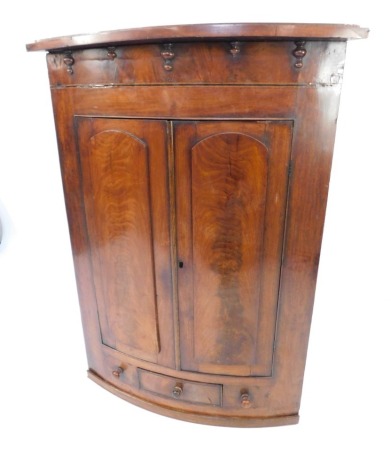 A 19thC mahogany bow fronted corner cabinet, the cornice with drop finials above two panelled and arched doors, with arrangement of one door flanked by two false drawers, each with knop handles, 124cm high, 90cm wide, 50cm deep.