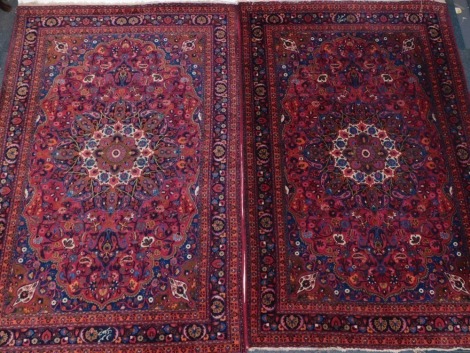 A pair of Persian rugs, each with central roundel and decorated with flowers, on a red ground with multiple borders, 220cm x 137cm.