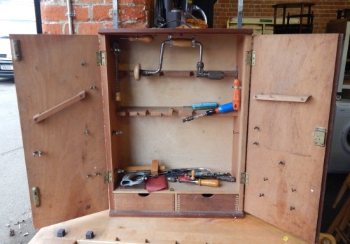 A pine tool cabinet, with two cupboard doors opening to reveal a fitted hanging tool interior, with a small selection of tools, including mainly screw drivers.