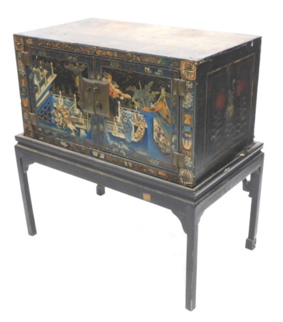 An antique Chinese lacquered chest on stand, decorated with figures on horse back, buildings, etc., on a black ground, with two doors enclosing a red painted vacant interior on a later ebonised base, 94cm high, the cabinet 85cm wide.