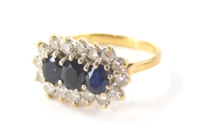 An 18ct gold sapphire and diamond cluster ring, the panel set with three oval cut sapphires, totalling approx 1.38cts overall, in a white gold claw setting surrounded by a halo of sixteen round brilliant cut diamonds, totalling approx 0.48cts, each in a c