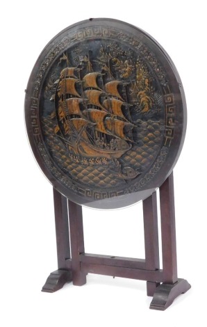 A mid 20thC Chinese tilt top occasional table, the circular top carved with a central ship in rough seas, glass protector, on an out folded base, 60cm diameter, 90cm high.