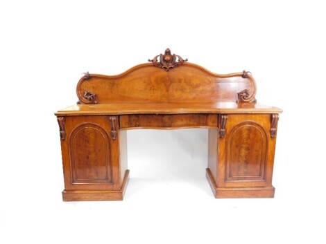 A Victorian serpentine mahogany breakfront sideboard, with a carved back, central cushion drawer, flanked by a pair of cupboard doors, opening to reveal a drawer and two slides to the left, a drawer and one deep wine cellar drawer, both raised on plinth b