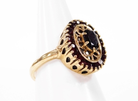 A garnet cluster ring, the central oval garnet in claw setting on raised section with outer design of tiny garnets, in a raised and pierced basket, yellow metal stamped 9ct, ring size O½, 4.9g all in.