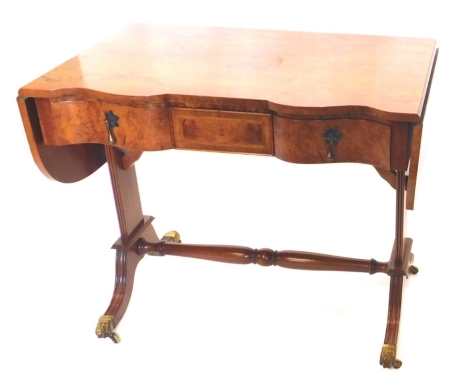 A walnut sofa table in the Regency style, the figured top with a cross banded border and a shaped front, above three feather banded frieze drawers, two with drop handles, on end supports with turned stretcher and reeded spade feet, terminating in paw cast