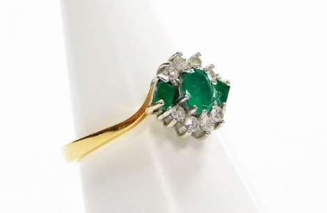 An 18ct gold emerald and diamond cross over ring, with three emeralds, one round brilliant cut and two baguette cuts, and eight round brilliant cut diamonds each in white gold claw stetting, on a yellow metal band, ring size M, 3.6g all in.