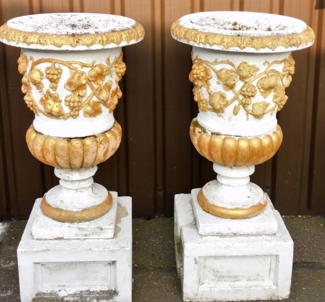 A pair of composition garden urns, each with a band of grapes and vines, on a tapering base with associated panelled plinth, later painted white and gilt, (AF), 114cm high.