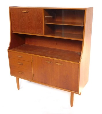 A 1970s teak sideboard, the drop down flap opening to reveal a drinks cabinet, flanked to the right with a pair of glazed doors enclosing a single shelf, above a pair of cupboard doors, flanked to the left by three graduated drawers raised on turned legs,