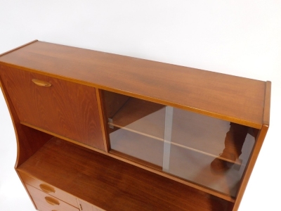 A 1970s teak sideboard, the drop down flap opening to reveal a drinks cabinet, flanked to the right with a pair of glazed doors enclosing a single shelf, above a pair of cupboard doors, flanked to the left by three graduated drawers raised on turned legs, - 2