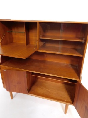 A 1970s teak sideboard, the drop down flap opening to reveal a drinks cabinet, flanked to the right with a pair of glazed doors enclosing a single shelf, above a pair of cupboard doors, flanked to the left by three graduated drawers raised on turned legs, - 3