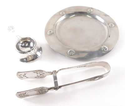 A Liberty Tudric pewter plate, decorated with roundels, impressed marks underside, 24cm diameter, a pair of silver plated asparagus tongs, and a silver plated tea strainer and stand.