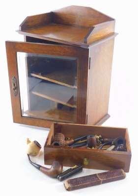An Edwardian oak smoker's cabinet, with three part galleried top, a glazed front door revealing fitted interior on plain base, with single drawer containing a quantity of various pipes and related items, Bass bottle cigar cutter, desk seal, cigarette hold