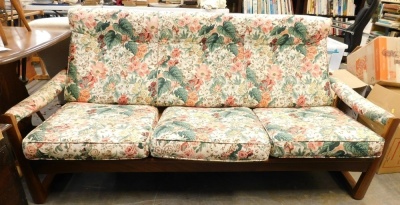 A 1960's teak Guy Rogers Virginia style three seater sofa, with later floral cushions to the padded back armrest and seat, 183cm wide. The upholstery in this lot does not comply with the 1988 (Fire & Fire Furnishing) Regulations, unless sold to a known e