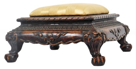 A stained and carved oriental footstool, with shaped top overstuffed in later Regency striped silk material, with an upper repeat floral banding, the border carved with further flowers, with dragon's mask corners, terminating in heavily carved ball and cl