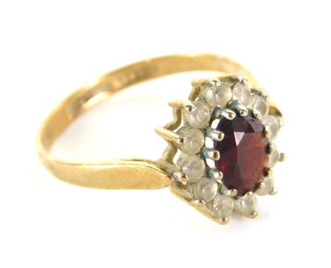 A dress ring, florally claw set with oval red stone, surrounded by small paste stones, part pierced shank marked 375, size O, 2g all in.