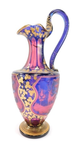 A late 19thC Bohemian pink and blue flashed glass ewer, decorated with shield reserves of deer and hunting dog, in a woodland setting, against a ground over decorated in gilt with trailing leaves, 33.5cm high. (AF)