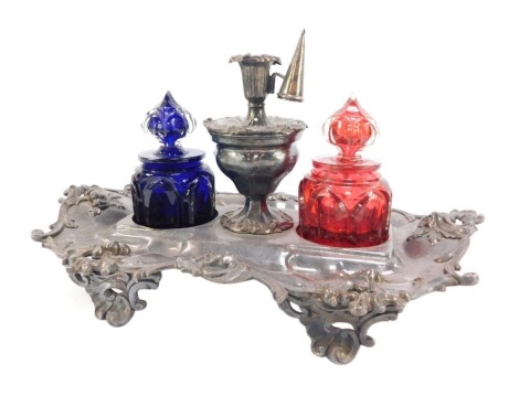 A Victorian silver plated desk stand, embossed with rococo scrolling leaves and dolphin's heads, with a central taper stick lidded mixing bowl, flanked by a pair of Gothic glass inkwells, one in cobalt blue, the other in ruby red, raised on scroll feet, 3