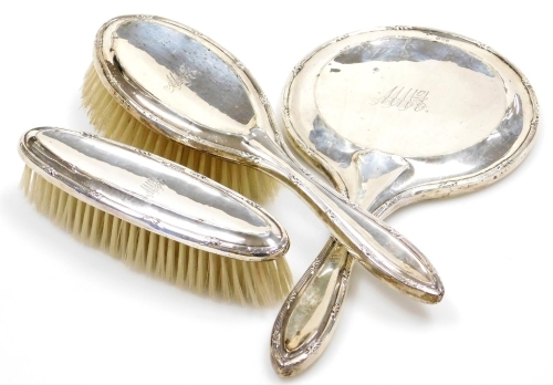 An Edward VII silver mounted three piece dressing table set, comprising mirror, hair brush and hand brush, each piece decorated with a banded and raised leaf border, monogrammed, Chester 1919.