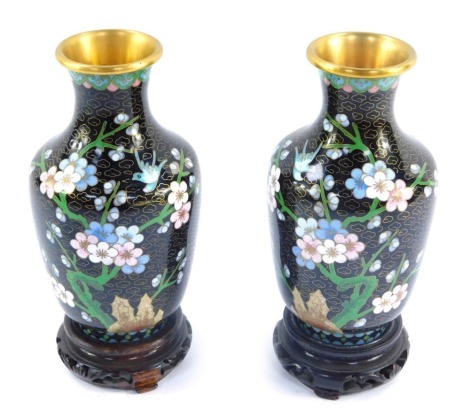 A pair of late 20thC cloisonne vases, on a black ground with blue, green and pink borders, with trees with floral sprays, on mahogany bases, the vases 15cm high.