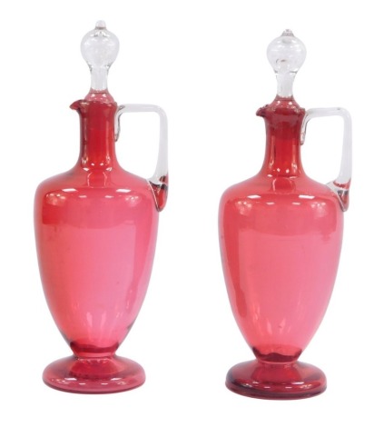 A pair of Victorian cranberry glass decanters and stoppers, the cranberry glass bodies with stepped foot and pouring handle, with later clear glass stoppers, 27cm high.