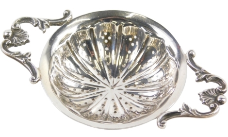 A George V silver tea strainer, the circular body flanked by scroll orb and shell handles, with pierced centre, Sheffield 1936, 11cm wide, 1.2oz.