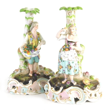 A pair of porcelain candlestick groups, formed as a lady and gentlemen, each dressed in finery, on naturalistic setting, set with flowers, in pink, green and blue, cross and T marks beneath, possibly Carl Theodore, c.1900, 19cm high. (2)