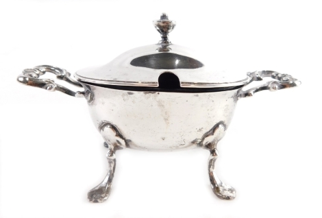 An Edwardian silver mustard pot, of oval form with scroll handles, on quadruple hoof feet with urn finial, hinged lid and blue glass liner, London 1901, 7cm high, 4.4oz all in.