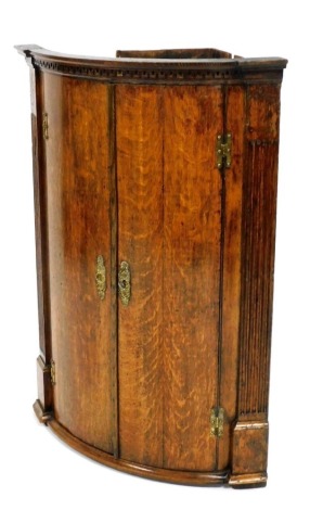 An early 19thC oak bow fronted hanging corner cupboard, with an inverted break front dentil cornice above two doors flanked by reeded pilasters, 101cm high, 69cm wide.