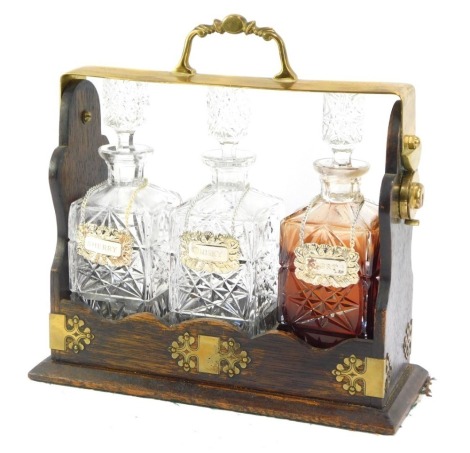 An early 20thC tantalus, with three cut glass decanters and three silver plated labels for sherry, whisky and port, in an oak case with brass bound supports, 36cm high, 36cm wide, 15cm deep.