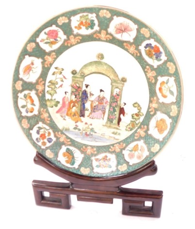 A late Chinese porcelain charger, decorated in coloured enamels with figures, etc, within a border of flowers and insects, on a green ground, 46cm diameter, with a hardwood stand.