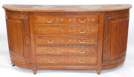 An Indian teak bow front sideboard, with floral carved and brass inlaid top, five graduated drawers to the centre enclosed by reeded curved doors, 91cm high, 190cm wide, 46cm deep.