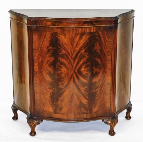 A reproduction mahogany and flame mahogany serpentine fronted side cabinet, the top with a carved and moulded edge, above a single door enclosing a single shelf, on four scroll capped pad feet, 102cm high, 103cm wide, 38cm deep.