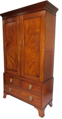 A George III flame mahogany linen press, with moulded cornice over two radiating flamed panel doors, enclosing an interior with an arrangement of press drawers and an optional hanging space, over two short and one long deep drawer, with heavy brass mask h