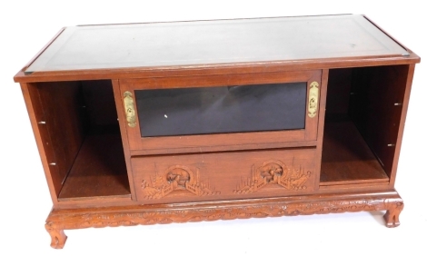 An Eastern hardwood television table, the panelled top with a glass inset above a door and a carved drawer flanked by two recesses, on shaped feet, 60cm high, 110cm wide.