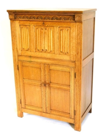 An oak cocktail cabinet, the frieze carved with arches above a linen fold carved fall front door with glass top and two panelled doors on stiles, 80cm wide.