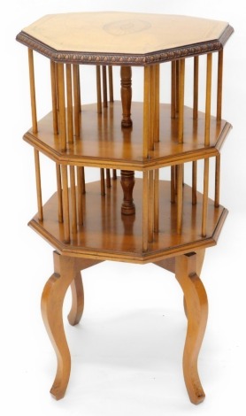 An Edwardian satinwood revolving bookcase, the octagonal top with a painted simulated inlaid oval patera, within a carved border, above two tiers for books, each with spindle turned dividers on splayed legs, 87cm high, 50cm wide. (AF)