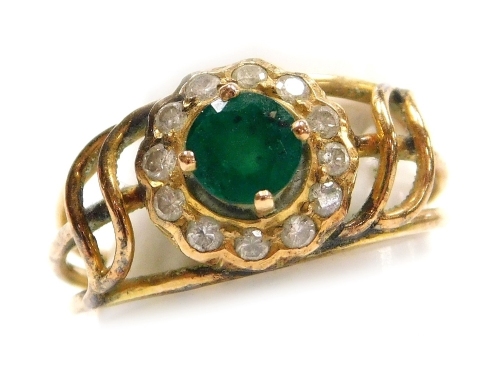 An emerald and diamond set dress ring, openwork design, a central round 4.9mm diameter emerald (heavily included) surrounded by twelve tiny diamonds set in yellow metal, size K, 3g all in.