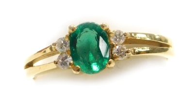 An emerald and diamond set dress ring, the oval faceted emerald of 5.2mm x 4mm with two tiny diamonds to each side, split shouldered shank, in yellow metal, size H, 2.2g all in.