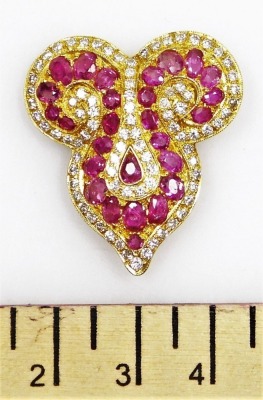 A ruby and diamond set brooch, of Eastern scroll shape set with twenty seven oval faceted rubies ranging in size from 1.3mm to 3mm long approx, with a tiny diamond set complete surround, the whole in precious yellow metal, pin back, 2.8cm x 107cm, 10.6g a - 2