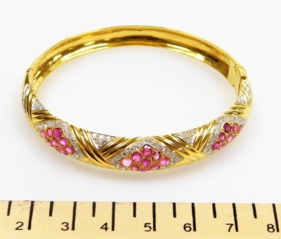 A ruby and diamond set hinged bangle, three ruby set lozenge shaped motifs of nine stones of 2mm each, surrounded by tiny diamonds with cross over raised pattern between, internal diameter 6cm, external diameter 6.5cm, 27.9g all in. - 2