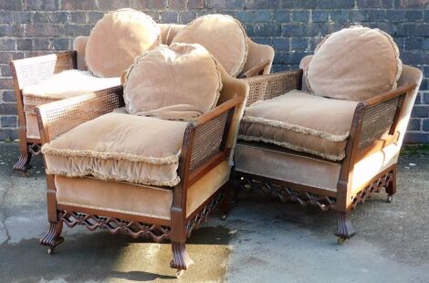 An early 20thC mahogany bergere suite, comprising two seater sofa, with cane filled arms and upholstered back and seat, with pink velvet fringed cushions, with a pierced apron scroll cast feet, and two matching armchairs, the sofa 136cm wide.